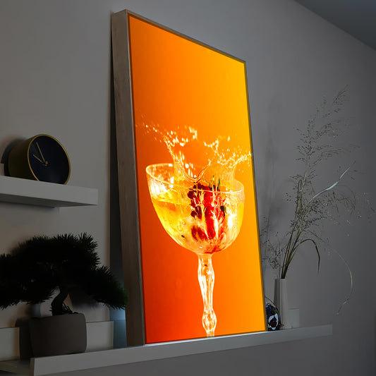 Gerahmtes LED-Bild mit Glas Orangensaft und Spritzern, schräger Blickwinkel
